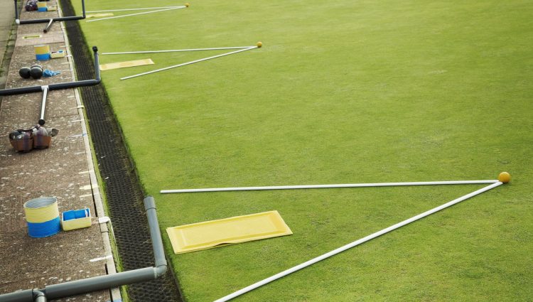 A lawn bowls green edge, a yellow player's mat to stand on and two white lengths of wood, guidelines with a yellow jack ball at the apex.